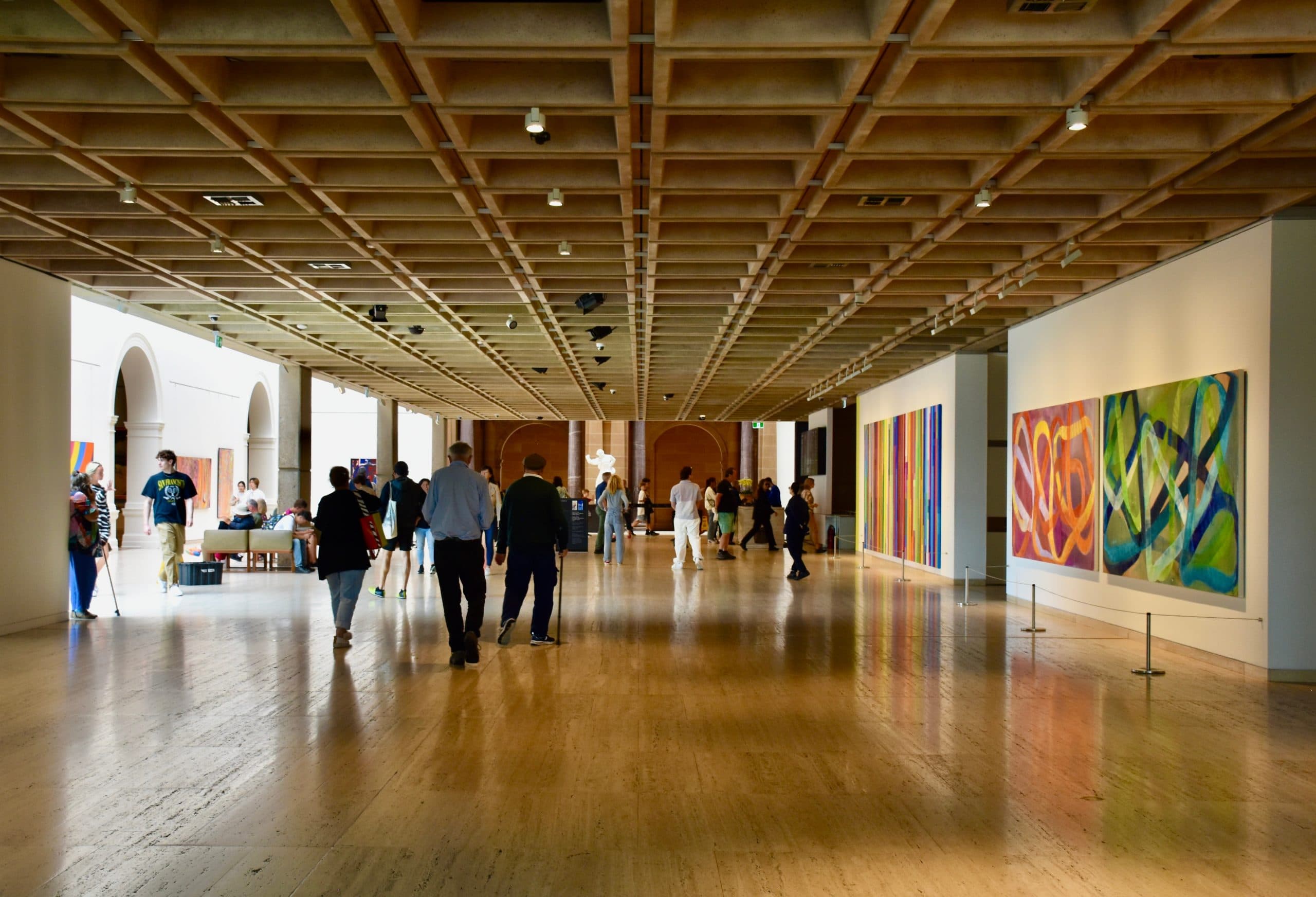 The hall on the main floor of Art Gallery of New South Wales, a large area with light brown timber floor and white walls. People are walking around looking at artworks on the walls.