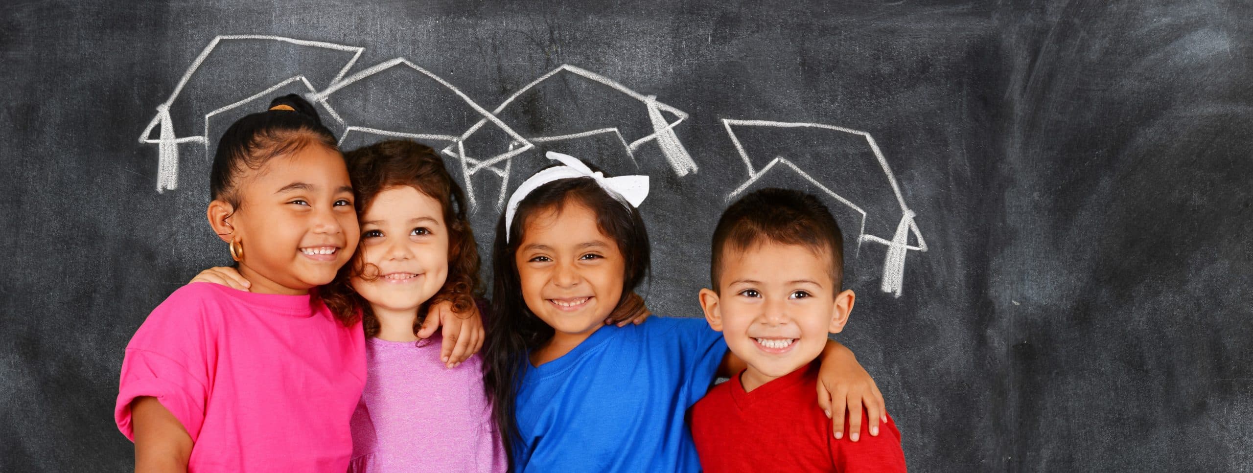 Four happy primary school aged children have their arms around each others shoulders. They are standing in front of a blackboard where graduate caps are drawn in chalk above their heads.