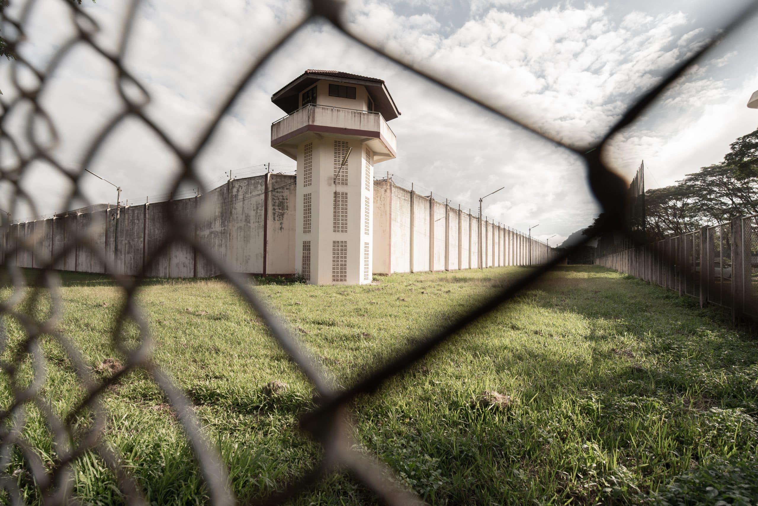Outside walls and towers of a prison, seen at a distance across an area of grass and through wire fencing.