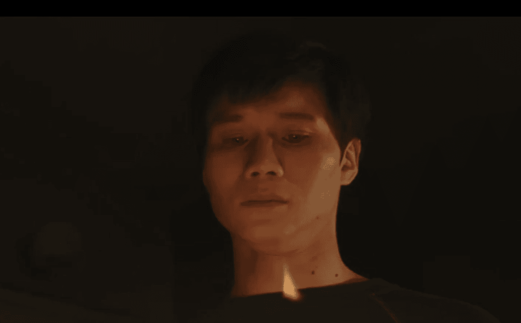 Actor Hoa Xuande playing The Captain in tv mini-series The Sympathizer. A man with dark hair standing alone against a darkened room above a flame.