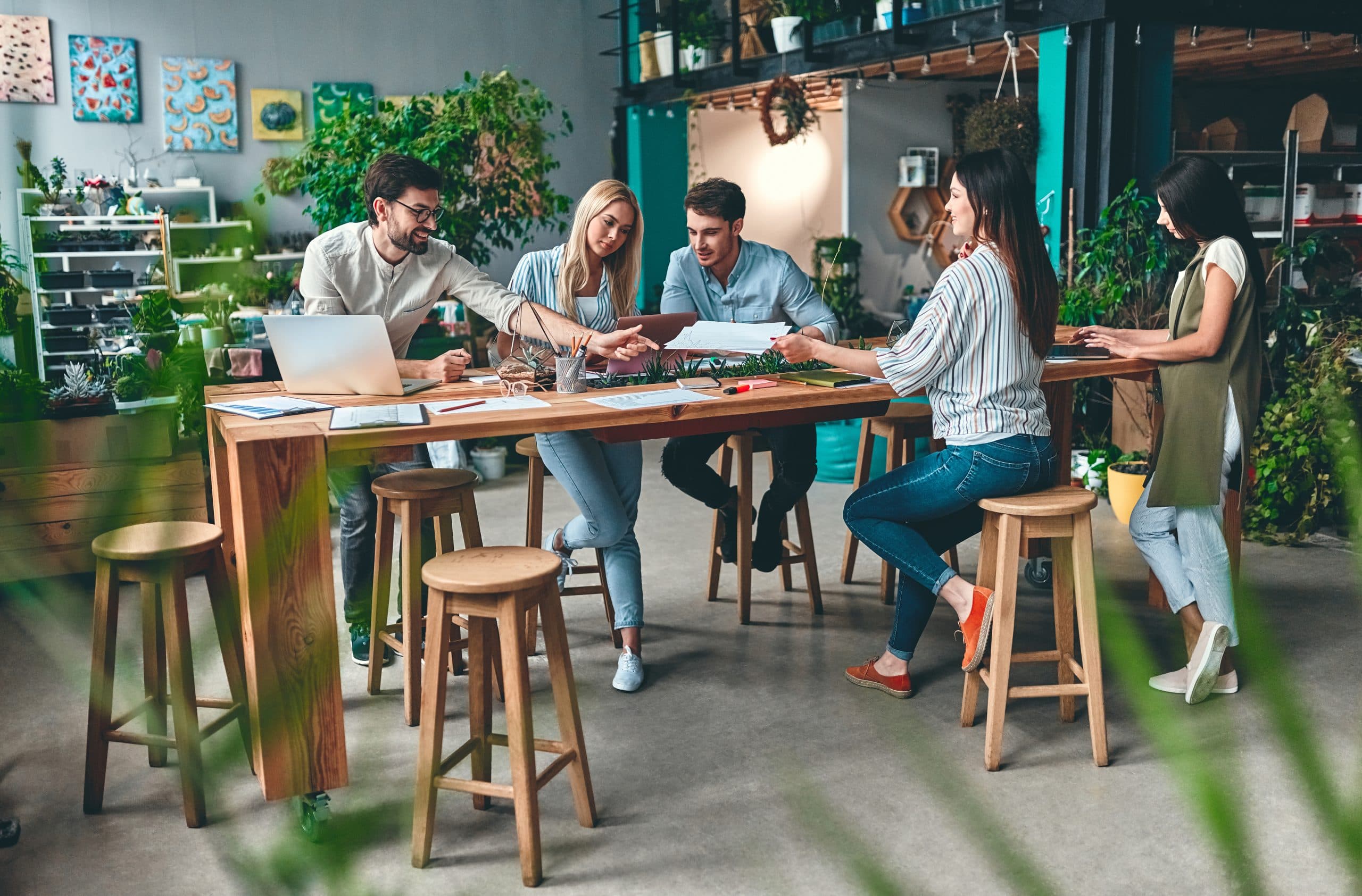 A group of young people in casual clothes are working together in a open workspace at a high desk. Shelves and plants are behind them.
