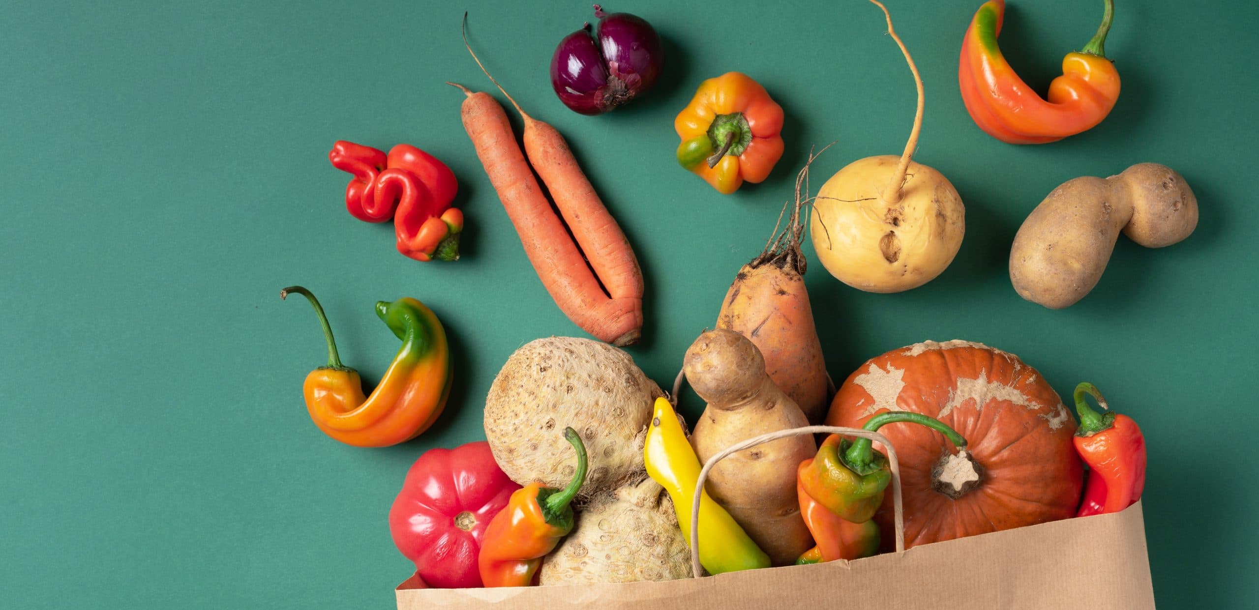Mix of misshapen fruit and vegetables spilling out of a paper bag, including carrots, potatoes and capsicums.