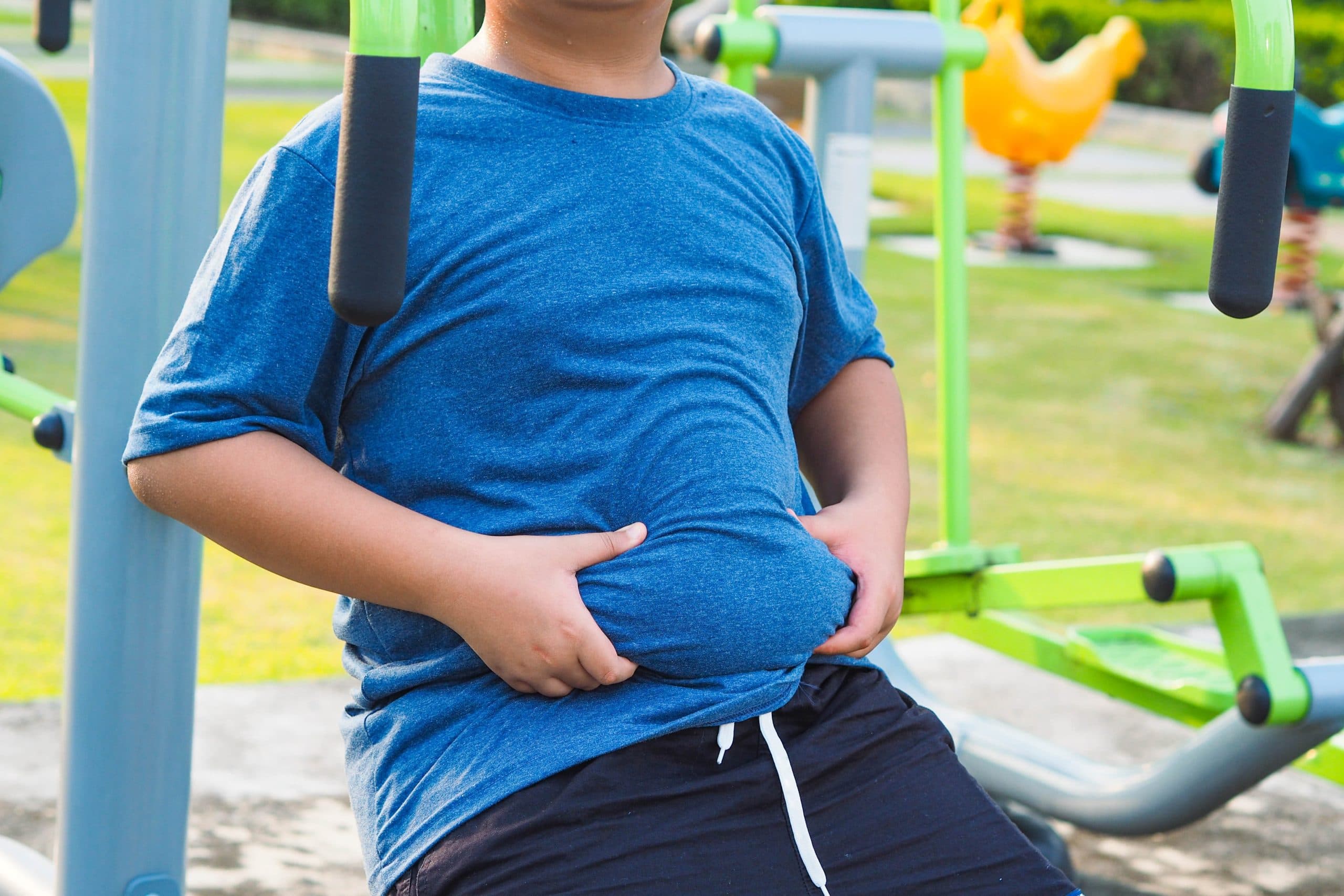 Zoomed in image of an overweight boy in bright blue tee shirt, so we can’t see his face. He is holding the bulge around his belly, while sitting in an outdoor kids’ playground.