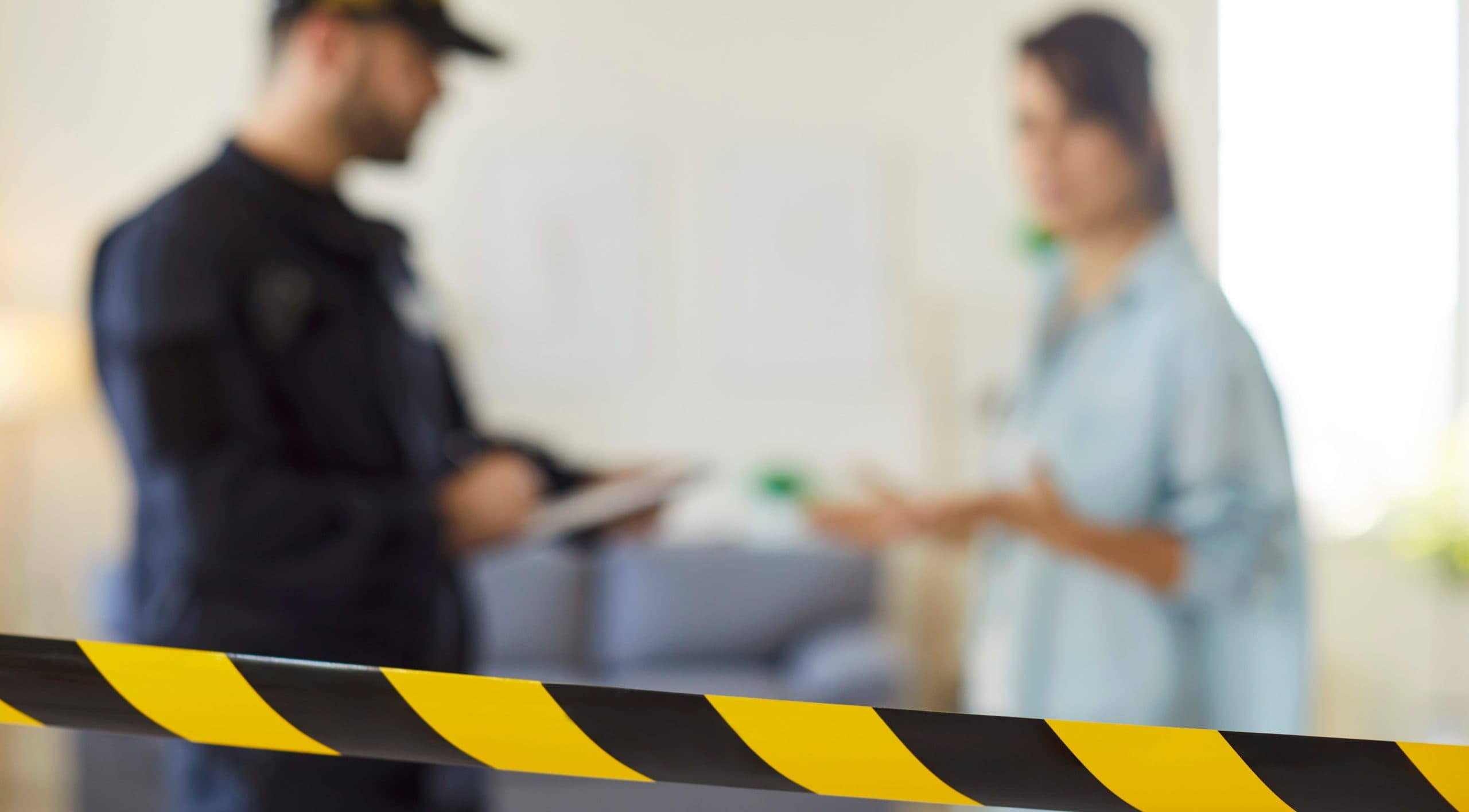 The scene of an incident in a room is taped off, and a woman in a blue shirt is standing talking with a police officer in a dark uniform taking notes.