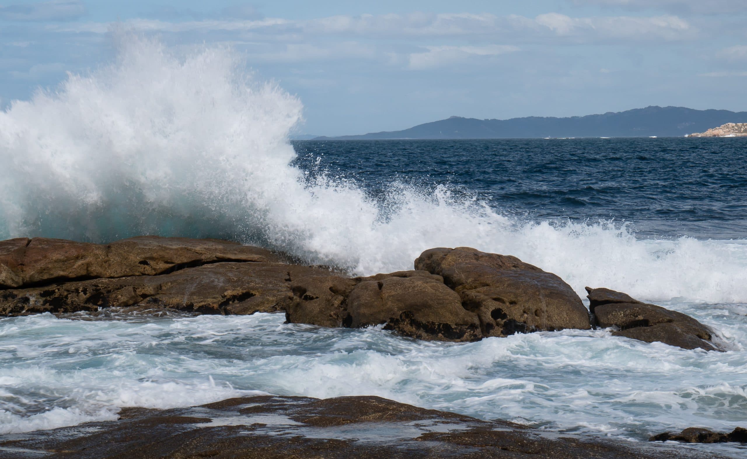 Fierce ocean waves breaking onto a rocky shore, with a coastline in the distance across a bay.