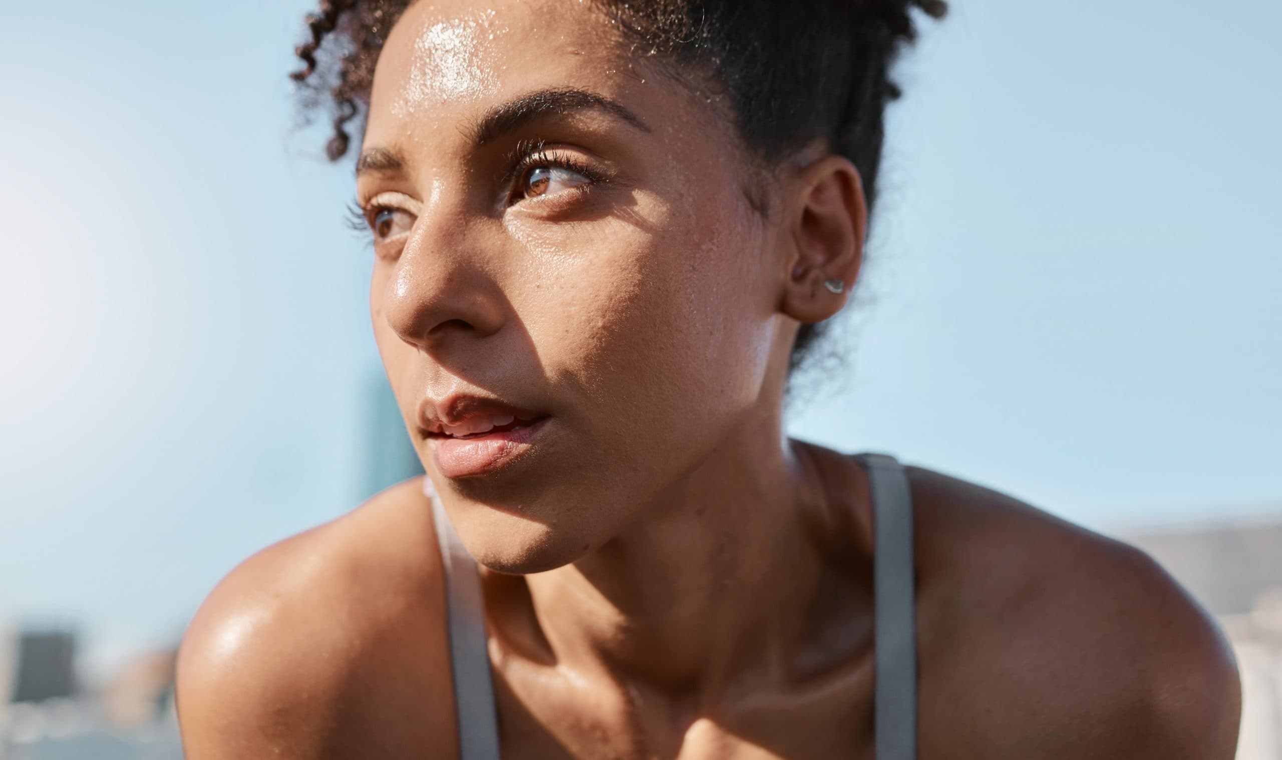 Head and shoulders of a young woman in a singlet who has been exercising outdoors, pausing to recover.