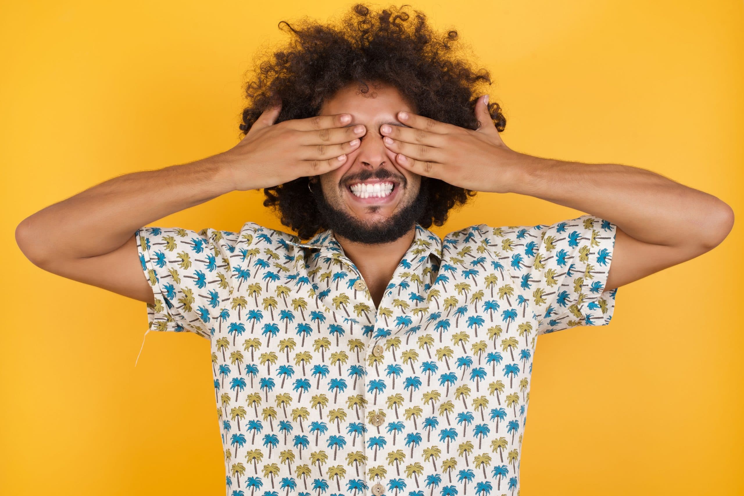 A young man wearing a bright patterned shirt covers his eyes with his hands while smiling.